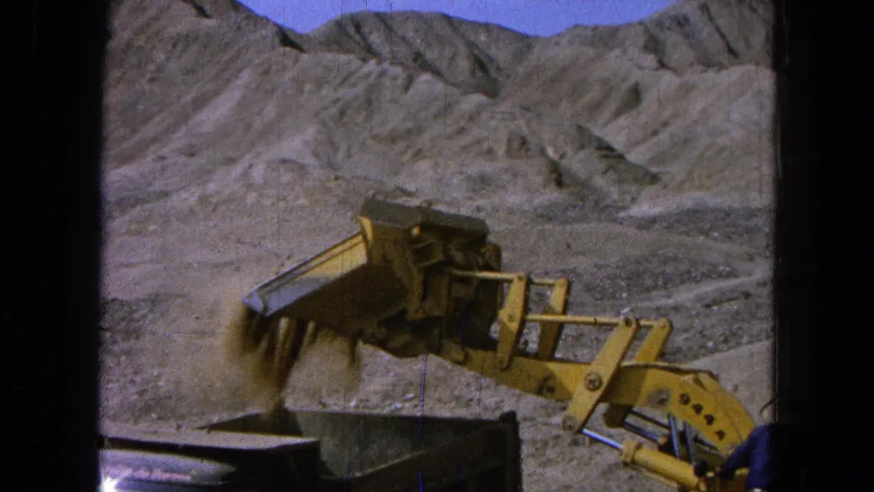 A yellow loader releases dirt into a dump truck during arid-site work in the 1965 Pueblo, Mexico footage.