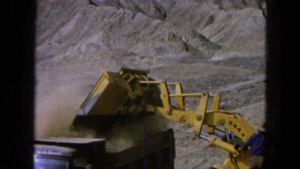Yellow loader works beside a truck in dusty terrain while a partly visible worker in blue stands near the machine.