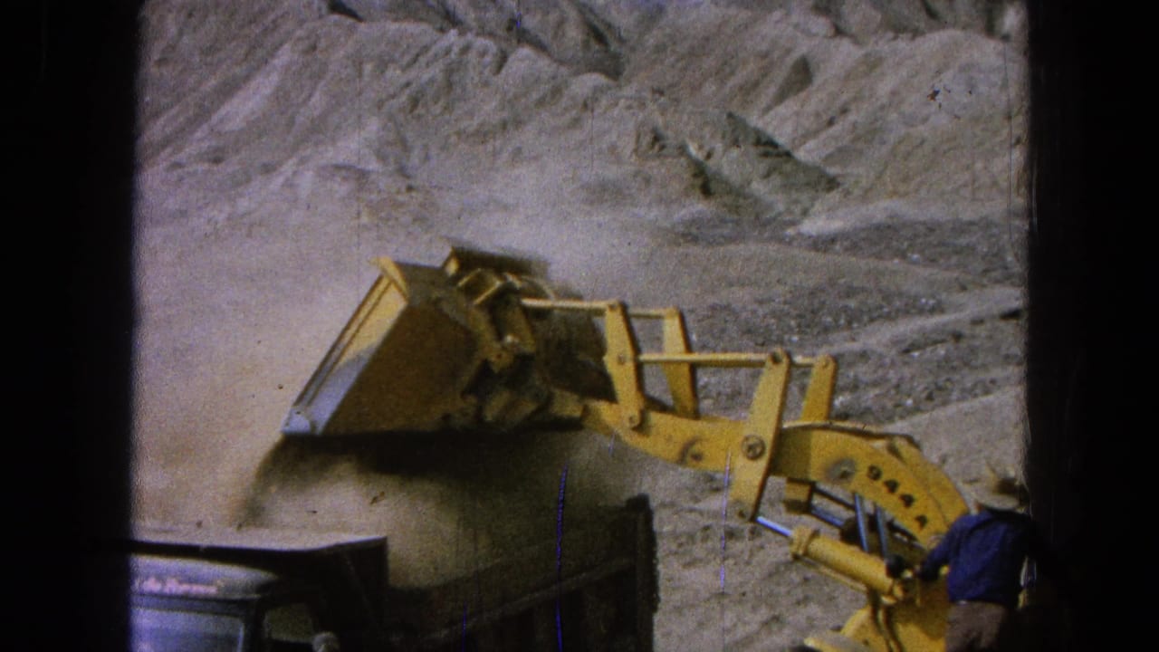 Yellow front-end loader dumping dirt into a dark truck bed at a dusty rocky worksite.