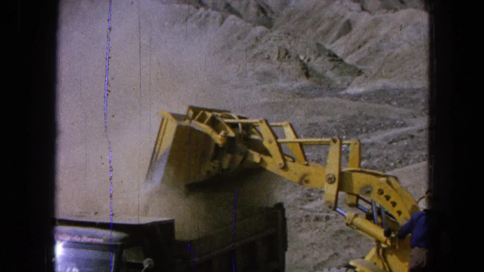 1965 Pueblo, Mexico 8mm frame showing the loader bucket angled over the truck bed as dust fills the left side of the frame.