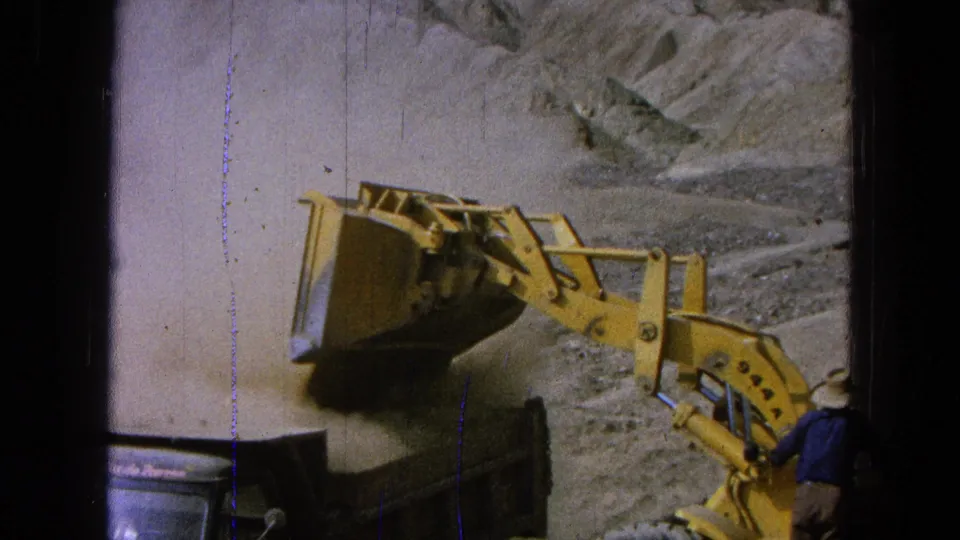 1965 Pueblo, Mexico 8mm frame with the yellow loader bucket raised after dumping into a truck, with dust and rocky terrain behind it.