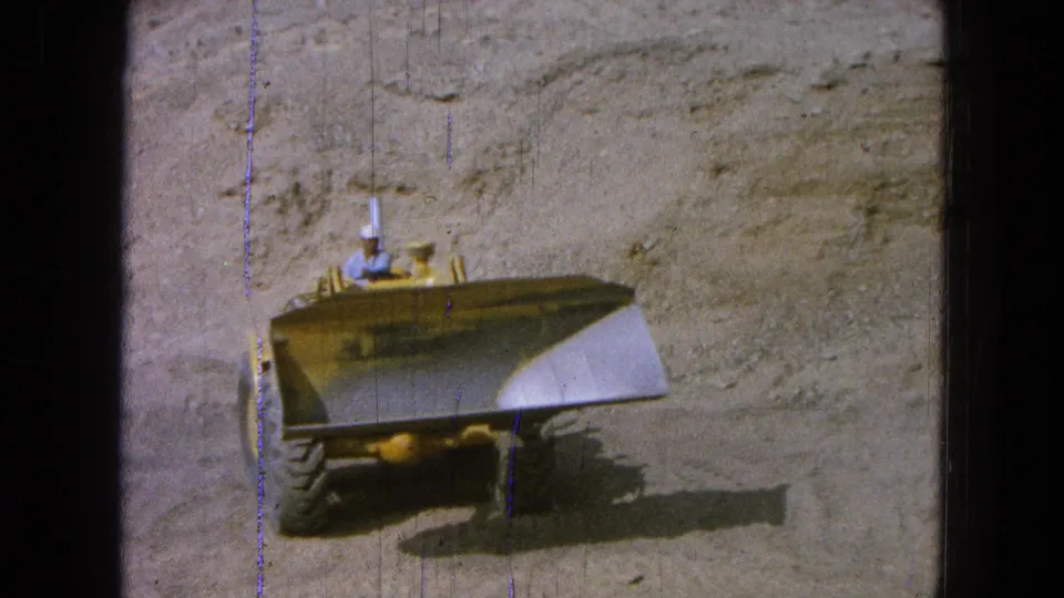 1965 Pueblo, Mexico 8mm frame showing a yellow front loader facing the camera on dusty ground with a worker seated at the controls.