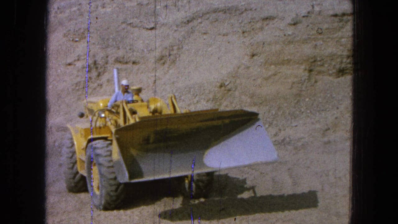 1965 Pueblo, Mexico frame of a yellow front loader shifting position beside a sandy cut, with the operator partly visible above the machine.