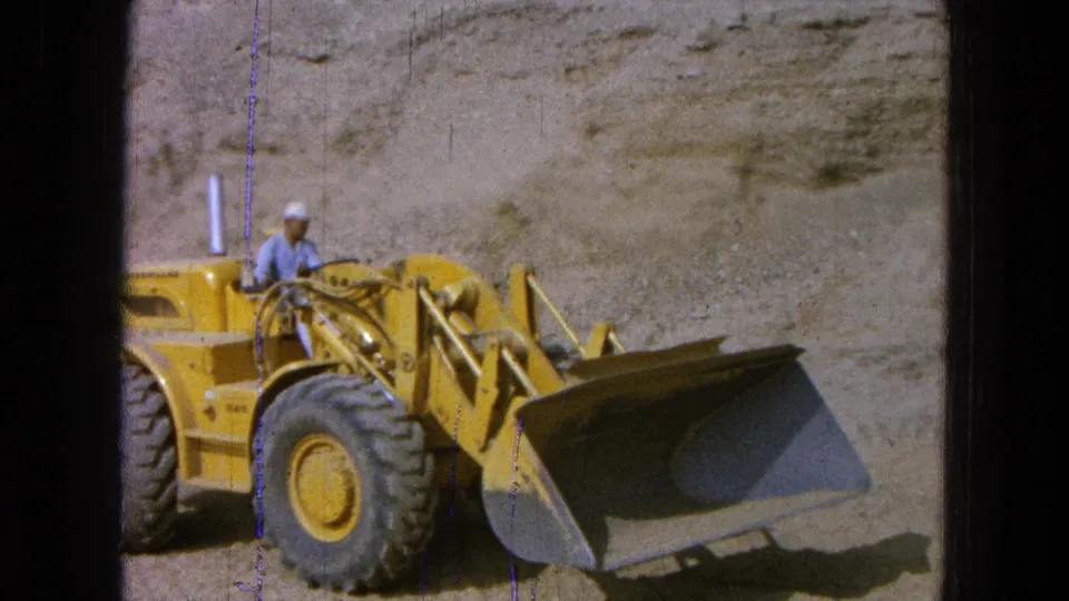 1965 Pueblo, Mexico 8mm still showing the side of a yellow front loader and its operator as the bucket extends across the dusty site.
