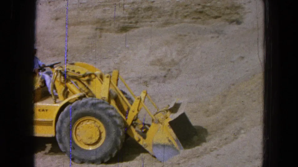 1965 Pueblo, Mexico frame showing the loader in side view as its bucket meets the dirt near a sandy excavation wall.
