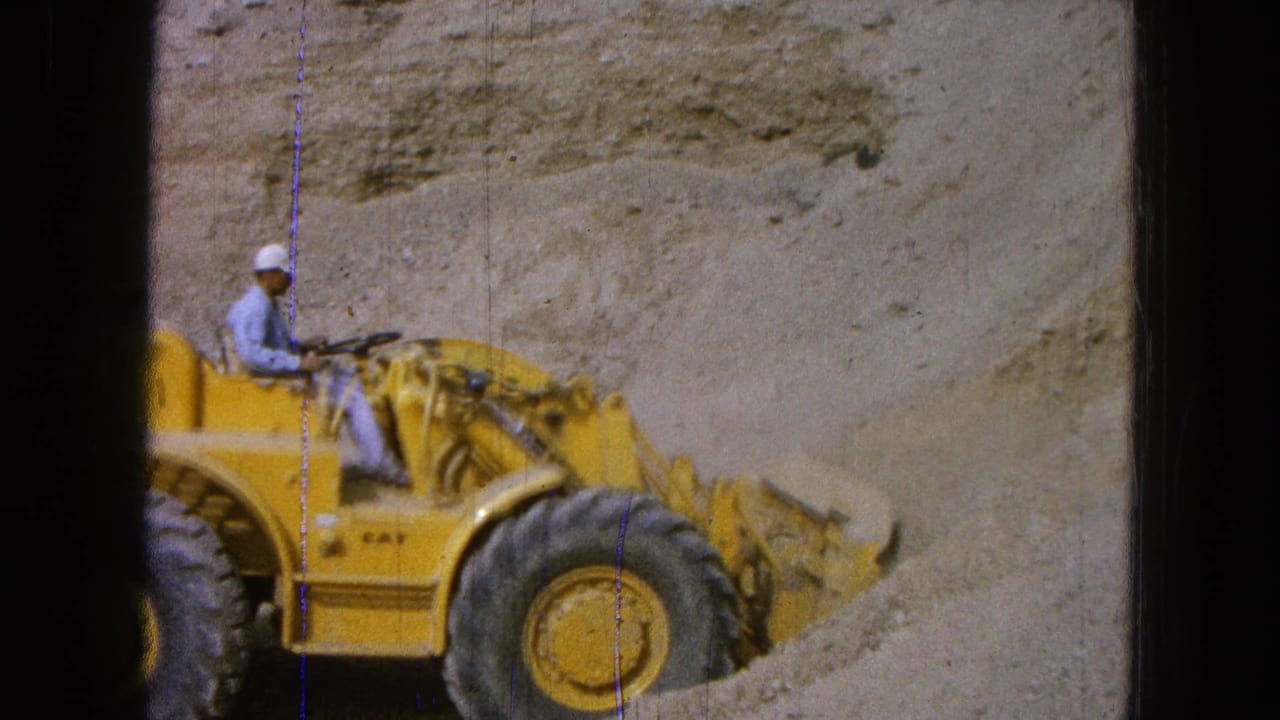 Worker in a white cap drives a yellow front-end loader along a sandy slope.
