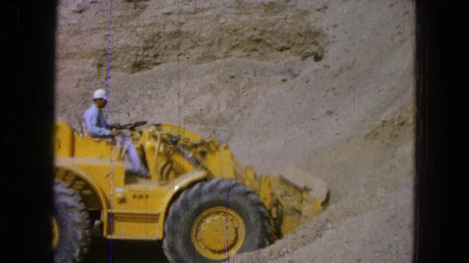 1965 Pueblo, Mexico frame showing a worker operating a yellow Caterpillar loader in a sandy construction area.