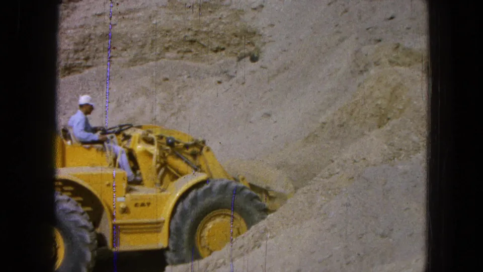 1965 Pueblo, Mexico frame with a seated worker driving a yellow loader beside a raised pile of sandy earth.