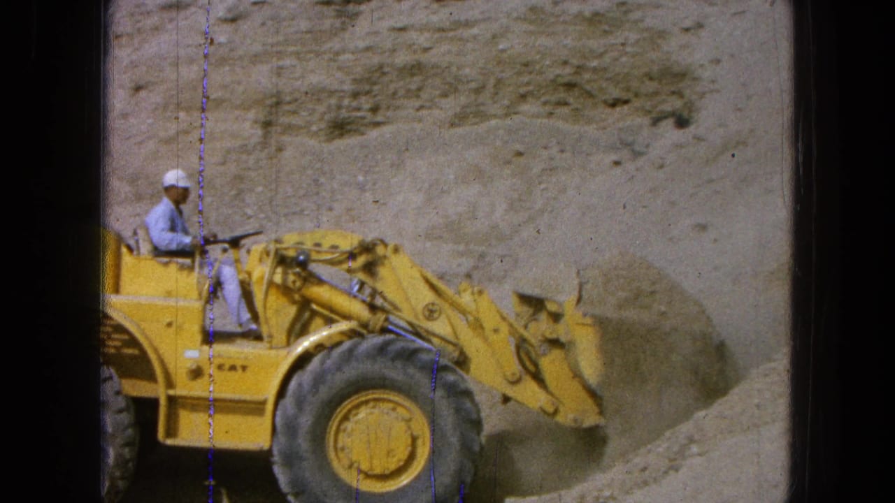 Worker driving a yellow front-end loader beside a sand pile at a dusty construction site.
