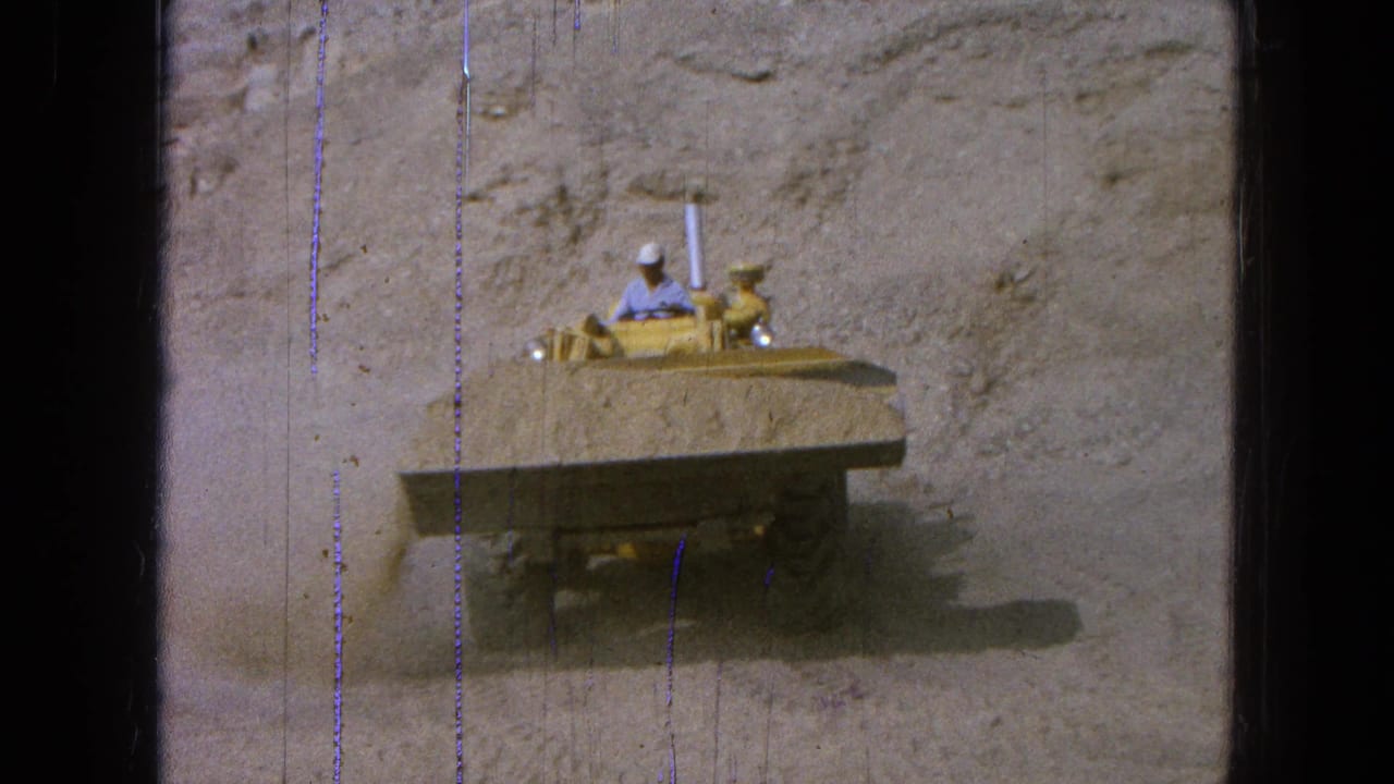 Yellow front-end loader drives across a dusty quarry floor with a helmeted operator visible.