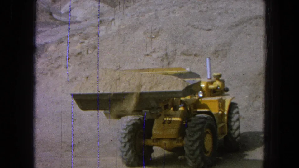 Yellow front-end loader turns with a raised bucket, heavy tires, and visible film wear in this 1965 Pueblo, Mexico quarry frame.
