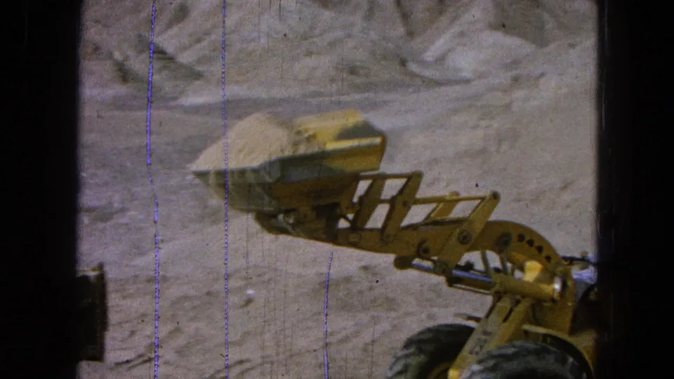 1965 8mm footage from Pueblo, Mexico shows a yellow front-end loader with a raised bucket of dirt at a dusty construction site.