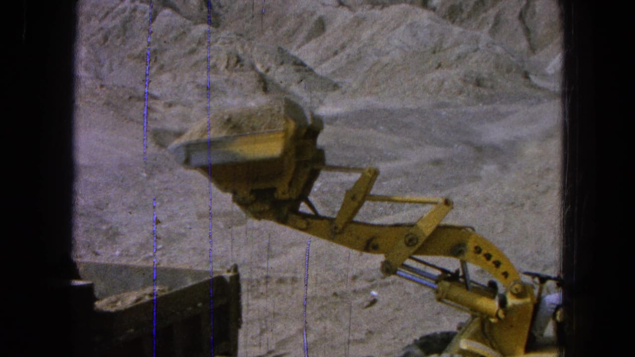 The loader arm extends over a dusty work area in Pueblo, Mexico, with the bucket approaching the dump truck bed.
