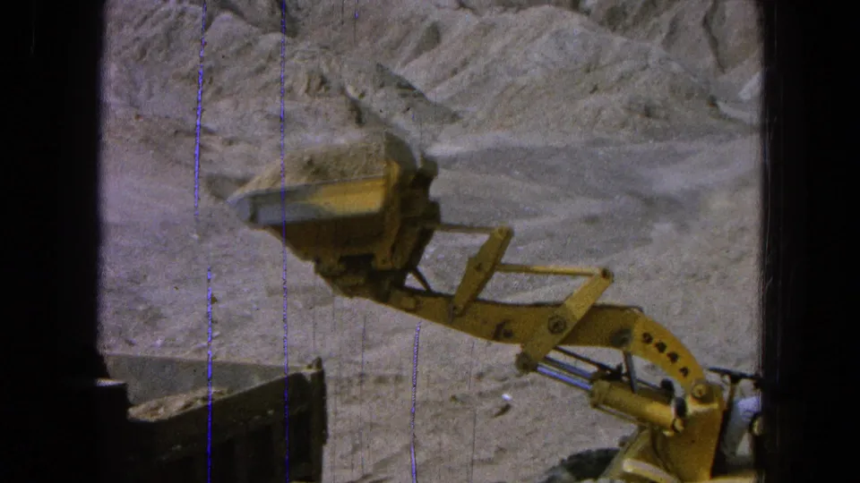 The loader arm extends over a dusty work area in Pueblo, Mexico, with the bucket approaching the dump truck bed.