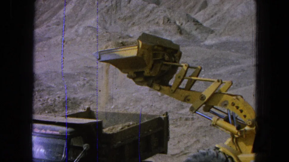 A yellow loader bucket is held over the dump truck bed, with exposed earth and 8mm film wear visible around the frame.