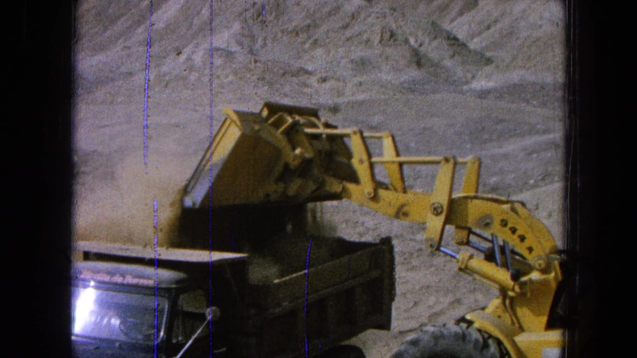 Yellow front-end loader raises a bucket beside a dark dump truck at a dirt worksite.