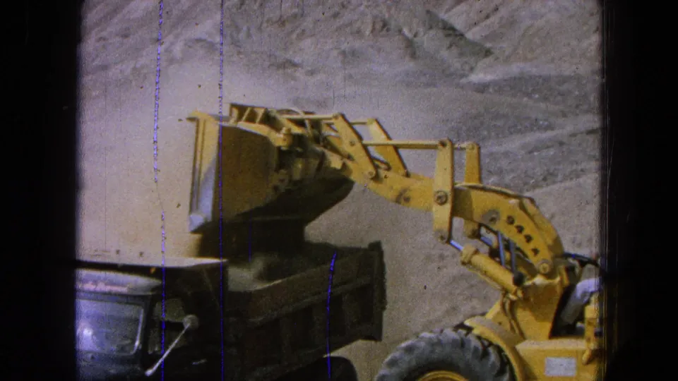 1965 8mm frame from Pueblo, Mexico showing a yellow loader bucket over a dark dump truck during earthmoving work.