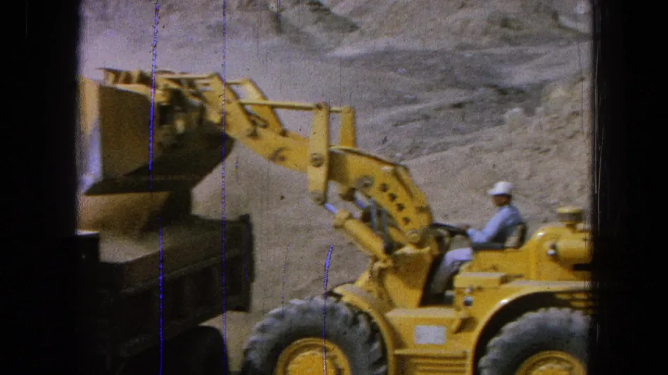 Hard-hatted worker operates a yellow front-end loader after dumping dirt near a dark truck in 1965 Pueblo, Mexico footage.