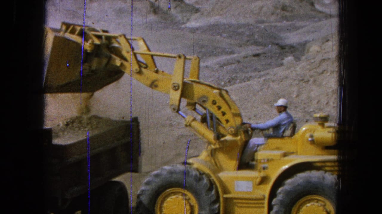 Yellow front-end loader beside a dump truck in a dusty quarry with an operator in a hard hat.
