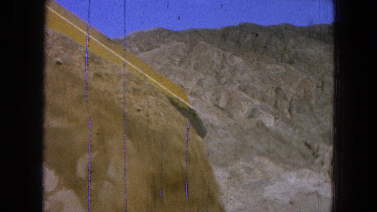Yellow loader bucket raised against rocky hills and blue sky.