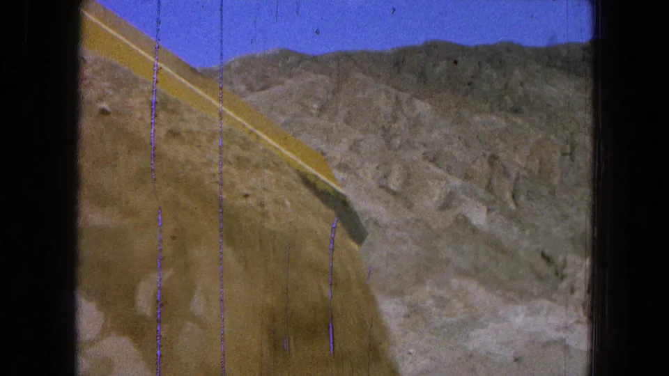 1965 Pueblo, Mexico 8mm frame showing a yellow loader bucket raised near rocky hills under a blue sky.
