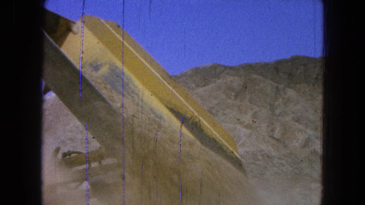 Raised yellow loader bucket with dirt and hills behind it.