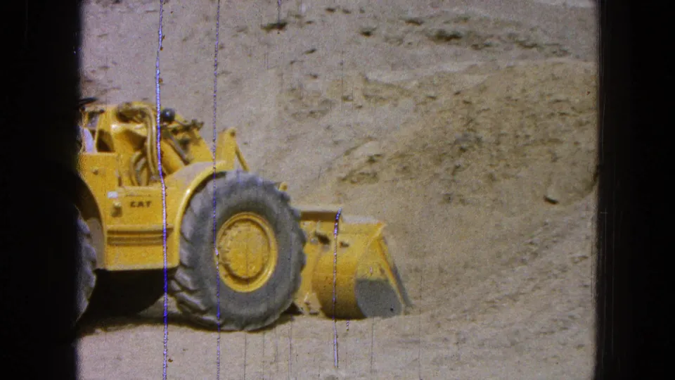 1965 Pueblo, Mexico 8mm frame showing a yellow Caterpillar front-end loader near a sloped dirt and gravel bank.