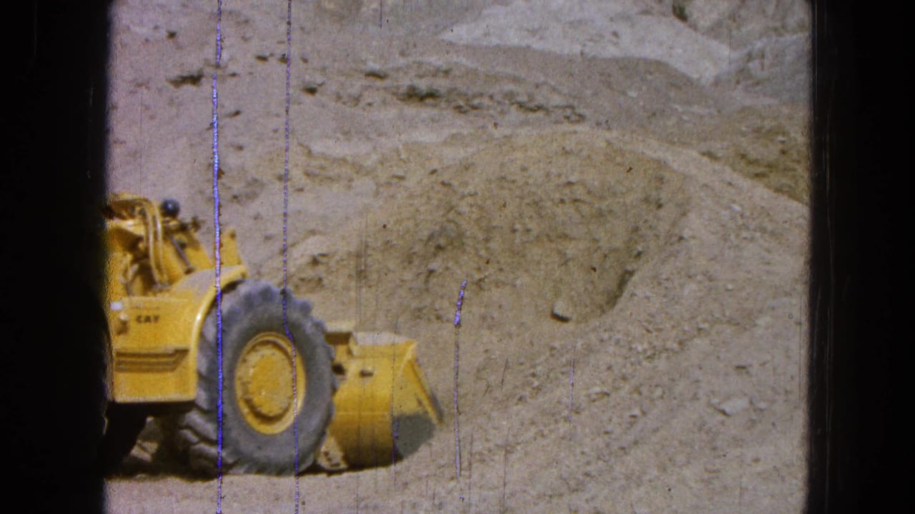 Yellow loader with its bucket near a gravel slope, partly cropped by the film frame.
