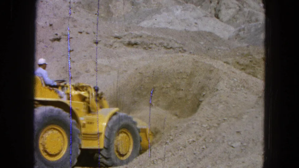 1965 Pueblo, Mexico 8mm frame showing a man in a white cap operating a yellow Caterpillar loader in a dusty work site.