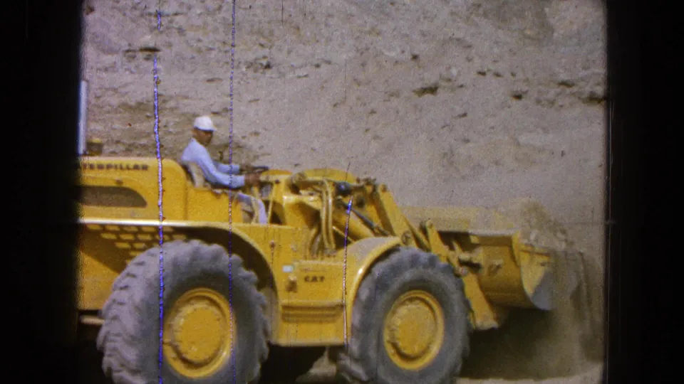 The operator sits at the controls of the yellow loader as the bucket carries or pushes sand in the dusty excavation area.
