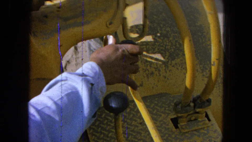 Handheld 8mm close-up of a hand near a black gear knob on yellow tractor controls, with worn floor plates visible.