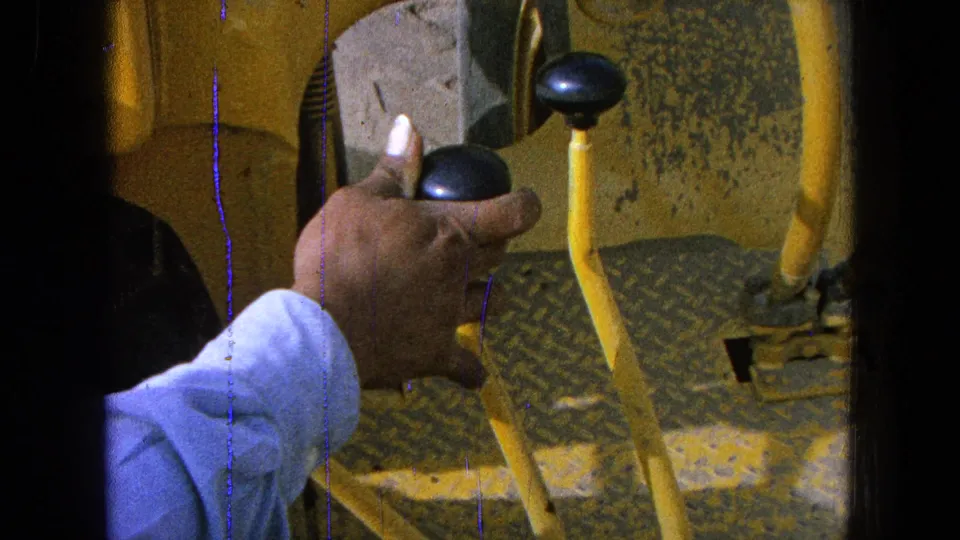 1965 home-movie frame showing a worker's hand holding a black knob on yellow industrial equipment in Pueblo, Mexico.