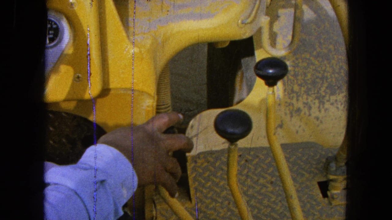 A hand grips a black control lever inside a yellow construction vehicle, with worn controls visible around it.