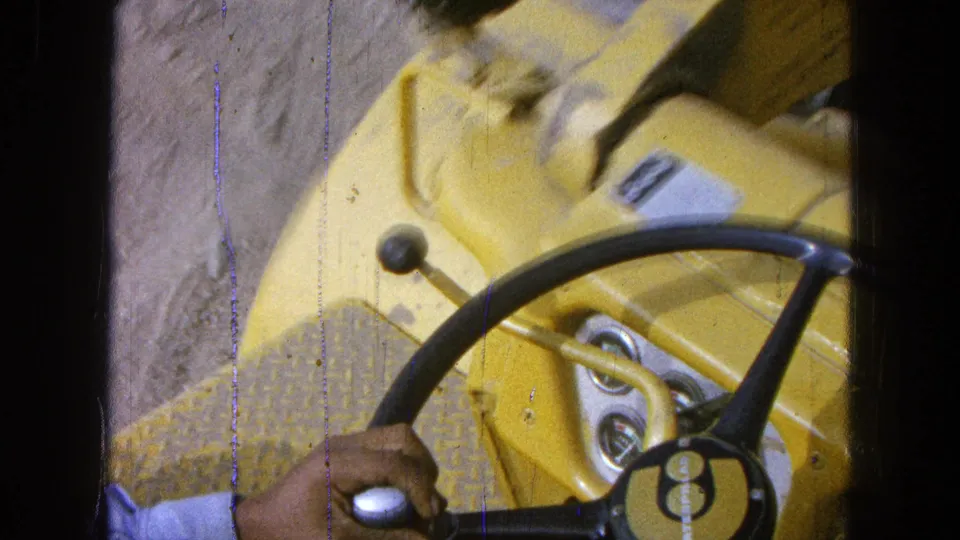 1965 Pueblo, Mexico 8mm frame showing a hand at the steering wheel and controls of yellow heavy equipment.