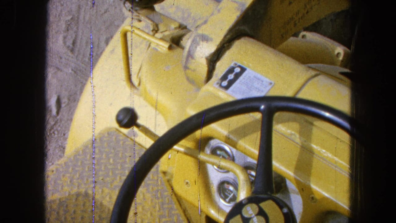 Yellow machinery dashboard with steering wheel, levers and gauges seen from above.