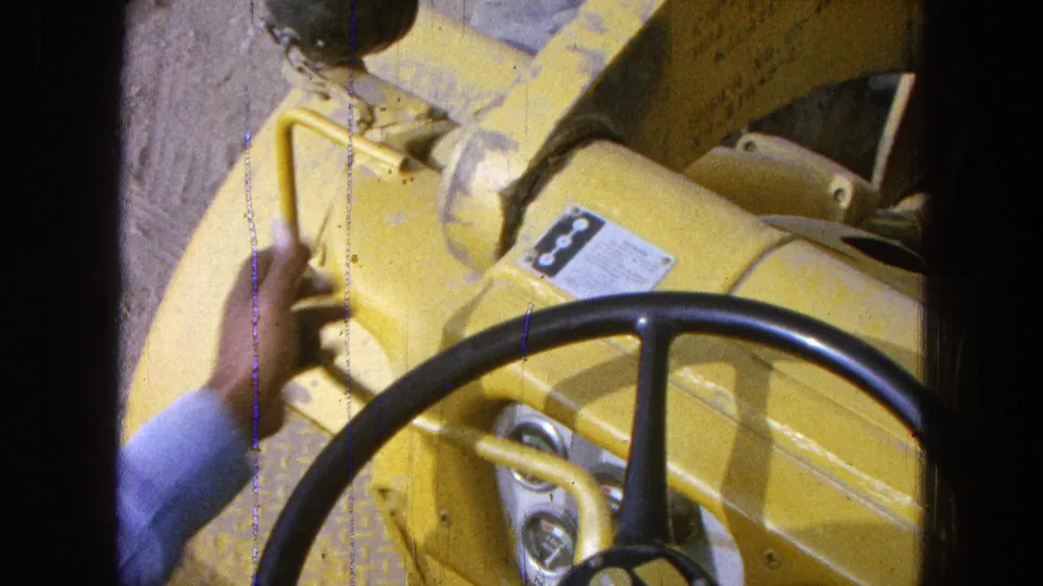 Operator's sleeved arm reaches near the lever and wheel of yellow construction or farm equipment in 1965 Pueblo, Mexico footage.