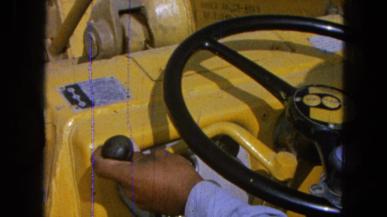 Hand on gear lever beside a black steering wheel inside a yellow farm machine.