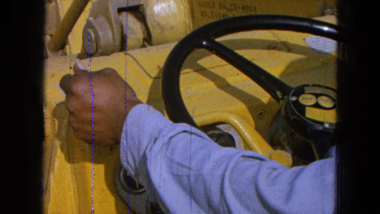 A blue-sleeved arm moves over yellow machinery controls in the 1965 Pueblo, Mexico farm-machine interior.