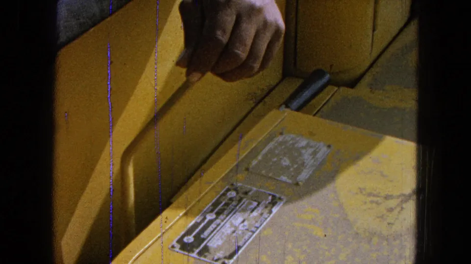 Close 8mm view of a hand pulling a slim control rod beside worn yellow panels and metal plates on farm machinery.