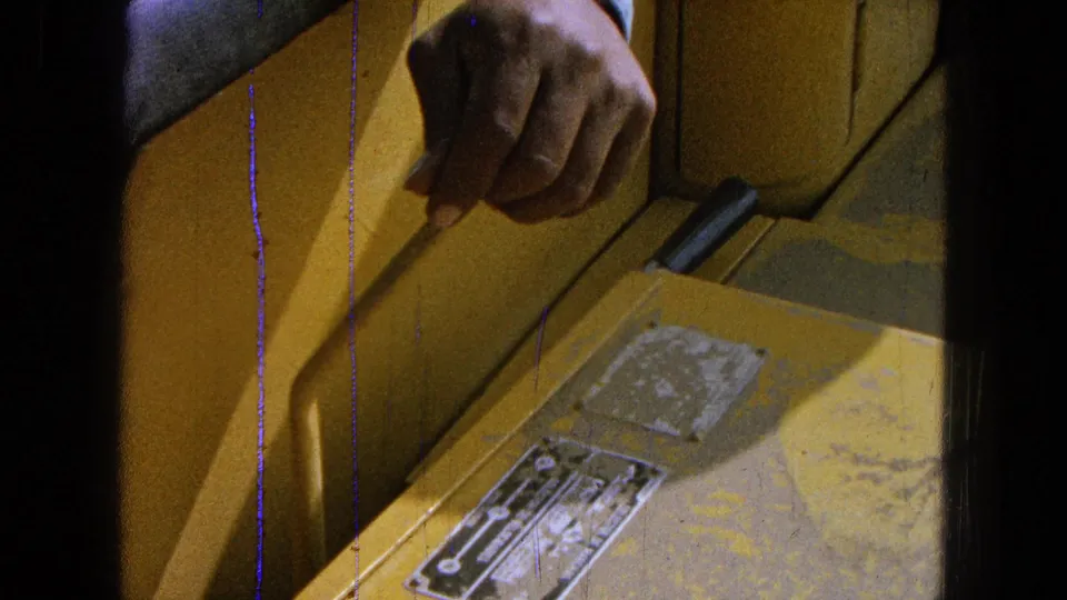 The operator's hand holds a side control inside the yellow farm machine, with worn plates and 8mm scratches visible.