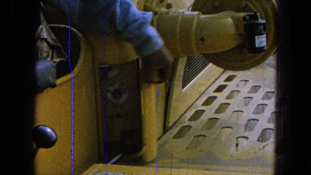 Close 8mm view from inside yellow machinery as a sleeved hand reaches toward a control area beside perforated flooring.