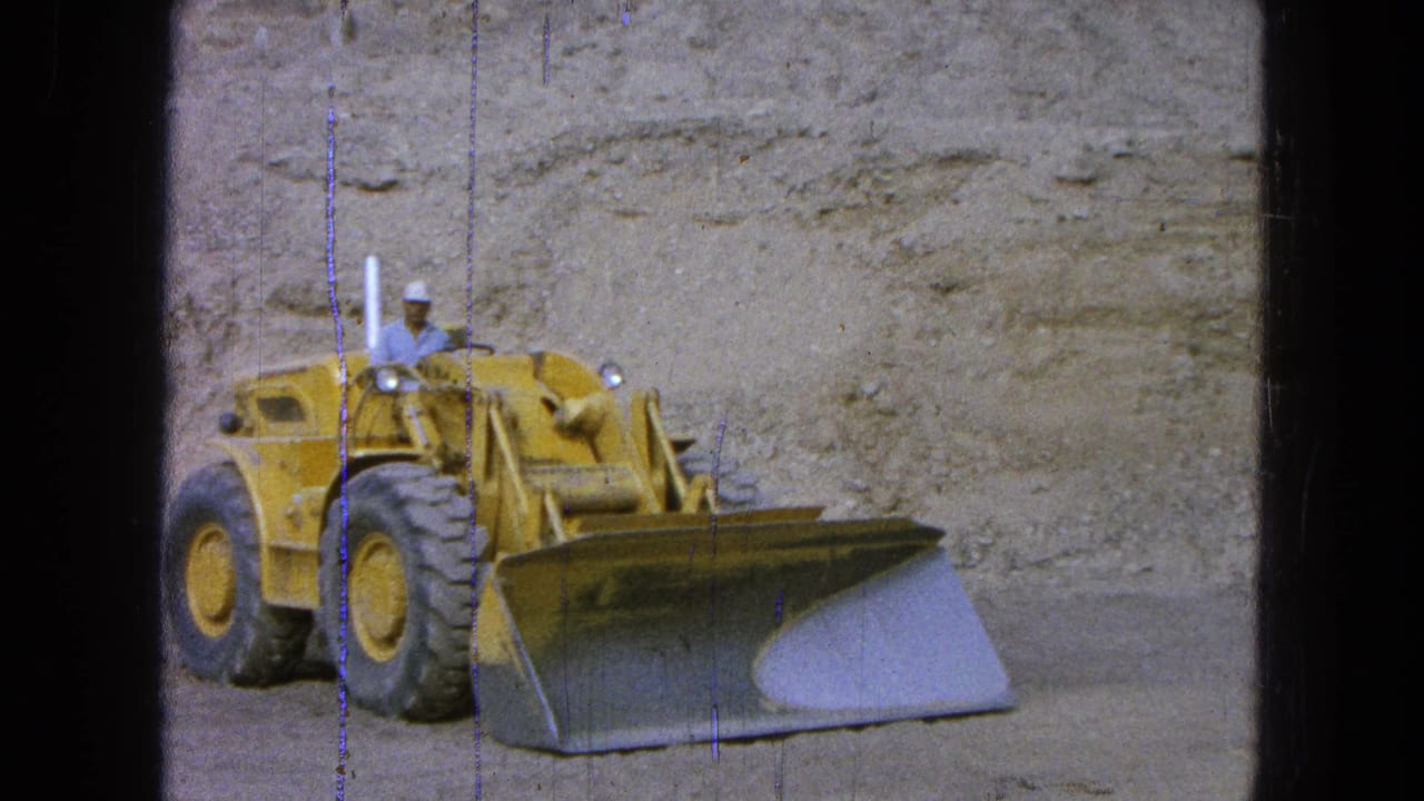 Yellow front-end loader with a hard-hatted operator beside a dirt embankment.