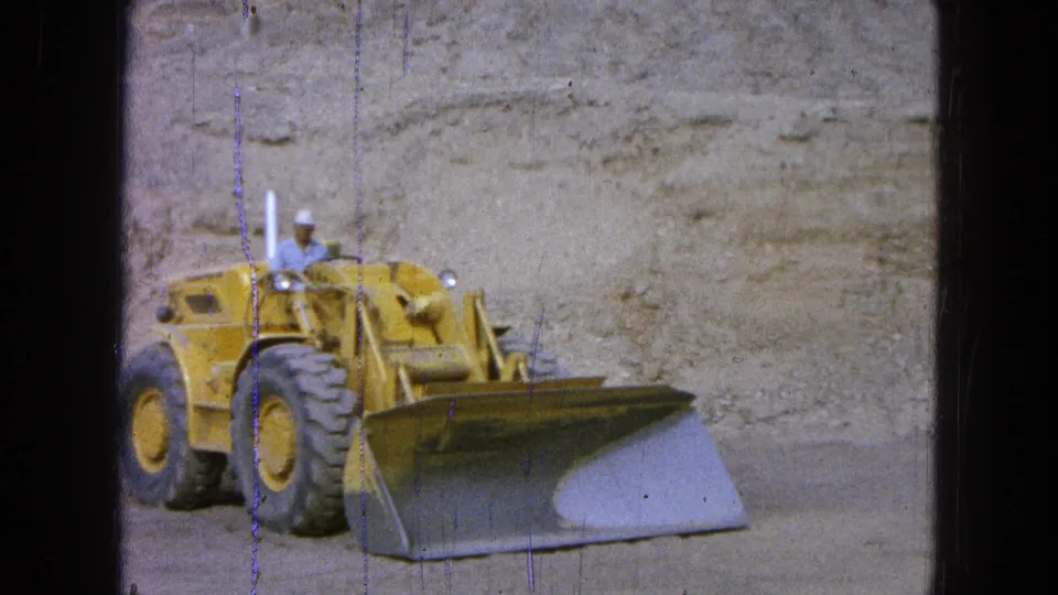 Side view of the yellow front-end loader in 1965 Pueblo Mexico, with the operator in the cab and bucket near the dirt surface.