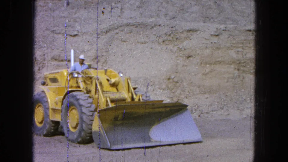 1965 Pueblo Mexico 8mm frame of a yellow loader facing forward, the hard-hatted operator seated behind the broad bucket.