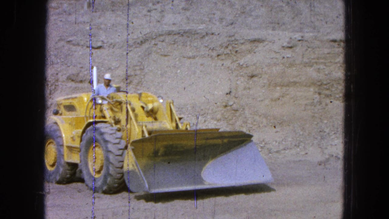 Yellow front-end loader with a hard-hatted operator in front of an excavated dirt wall.