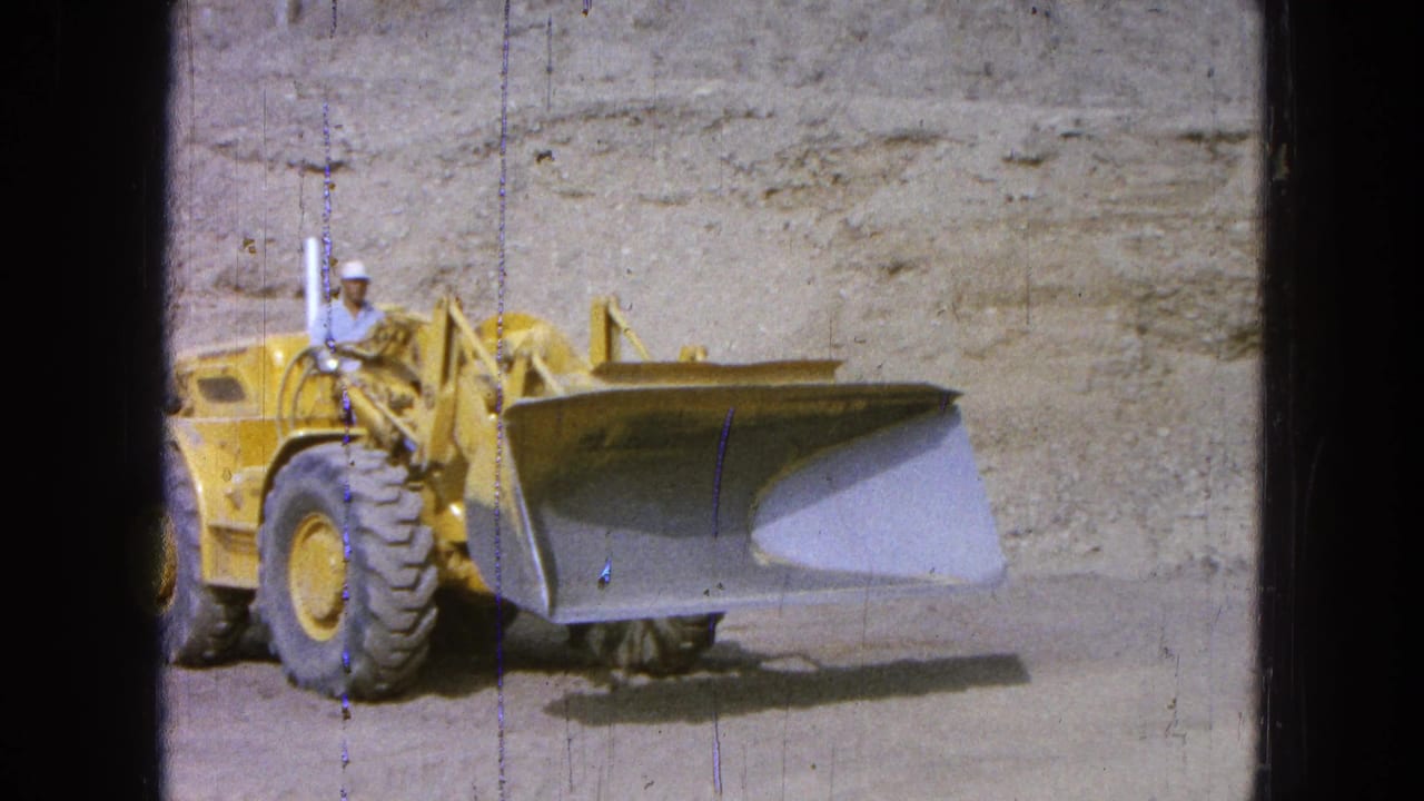 Yellow front-end loader with an operator approaches on dirt near an earthen wall.