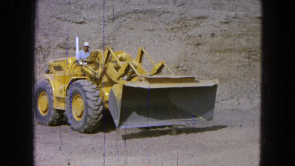 The 8mm frame shows the loader angled at the excavation site, with its broad bucket extended and the operator visible above the wheel.