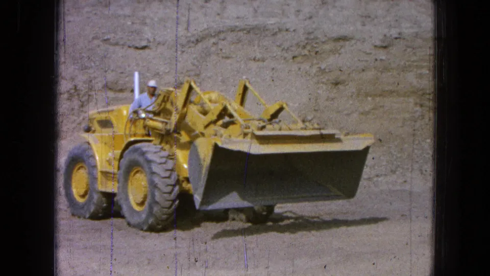 A hard-hatted operator sits on the yellow front-end loader as the lowered bucket faces the camera in the Pueblo, Mexico construction scene.