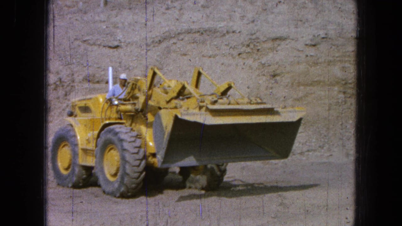 Yellow front-end loader with a hard-hat operator drives beside an earthen wall.
