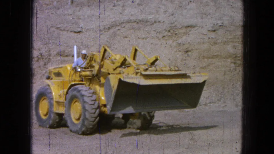 1965 Pueblo, Mexico 8mm frame showing a yellow front-end loader with a hard-hat operator near a dusty earthen wall.